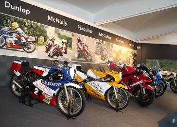 Five road racing motorbikes are on display in a line. On the wall behind them is the surname and photo of the person that road each bike 